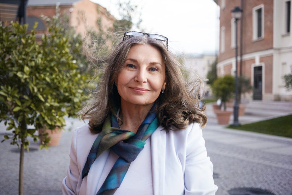 Outdoor image of gorgeous positive lady with charming smile and loose gray hair enjoying nice summer day, wearing white jacket, silk scarf and eyeglasses. Beauty, urban style and fashion concept